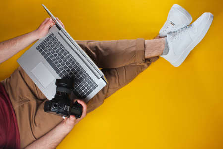 Conceptual photo of work photographer. Man sits on yellow background and holds laptop and camera. Top view. Minimalismの写真素材