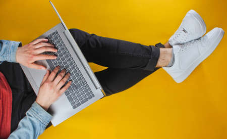 Online worker concept. Hipster man typing on laptop and sits on yellow background. Studio shot. Top viewの写真素材
