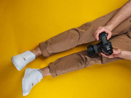 Man in beige pants and white sneakers holding a camera while sitting over yellow background. Top view. Studio shotの写真素材