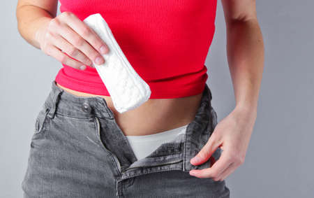 Woman with unbuttoned jeans holds pads in her hands on gray background. Menstrual days concept. Personal hygieneの写真素材