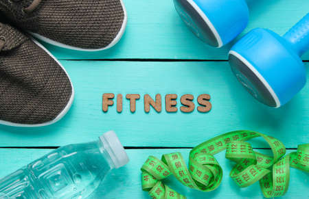 Healthy lifestyle. Dumbbells, ruler,  bottle of water, sneakers on blue wooden background with word fitness. Slimming concept.の写真素材