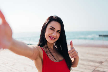 Young smiling woman in a red swimsuit takes a selfie on the beach