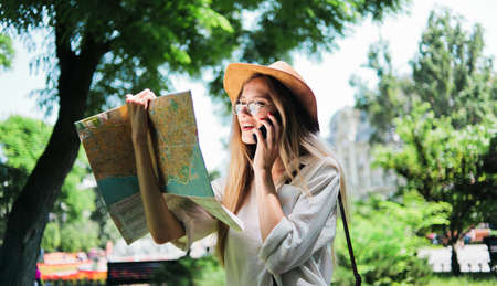 Tourism concept, travel to new cities. Attractive woman tourist in a felt hat and glasses looks into the city map and talks on the phone outdoor.の写真素材