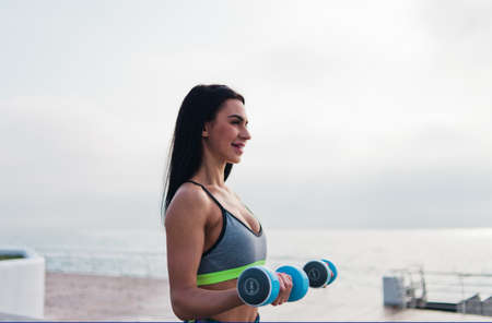 Young fit woman in sportswear doing exercise with dumbbells at the morning on sunrise beach