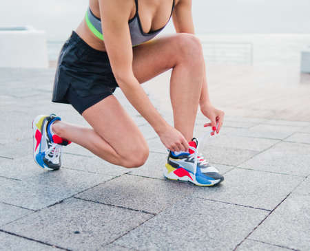 Fit woman in sportswear tying shoelace of sports shoes on a concrete tile and preparing to run on a...