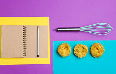 Minimalism Italian cuisine concept. Raw tagliatelle noodle, notebook recipe, whisk on colored paper backgroundの写真素材