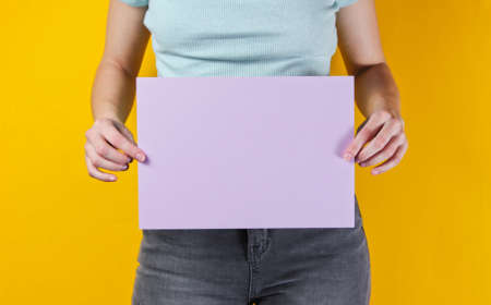 Woman holding purple empty sheet of paper for copy space on yellow background. Crop photo, studio...