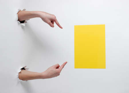 Female hands point fingers at yellow sheet of paper for copy space through the torn holes of white background. Creative artの写真素材