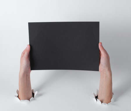 Female hands holding black sheet of paper for copy space through the torn holes of a white background. Creative artの写真素材