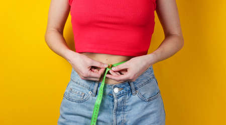 Weight loss concept. Woman tries on larger jeans and measures her waist with a ruler on a yellow backgroundの写真素材
