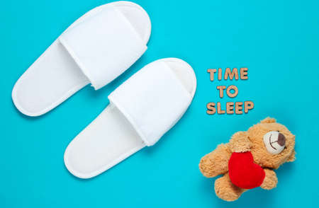 Time to sleep. White sleeping hotel slippers with a teddy bear on blue background. Top viewの写真素材