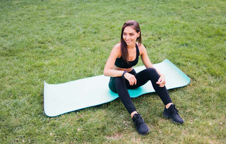 Cheerful healthy fitness woman in sportswear is sitting on a yoga mat and smilling in park on the...