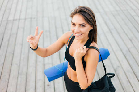 Slender cheerful fit woman in sport clothes with gym bag and mat on her shoulders posing after training outdoorsの写真素材