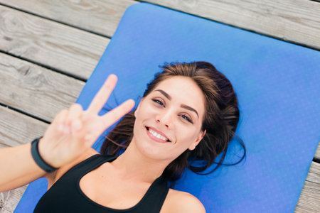 Young cheerful sport woman lies on mat and shows V symbol. Top view portraitの写真素材