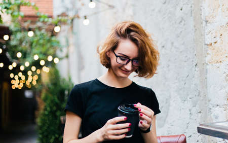 Cheerful hipster girl in glasses with short haircuts holding coffee cup against the background of...