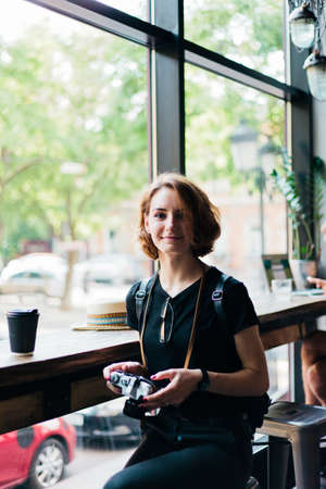 Young hipster woman holds retro camera in her hands and look at camera in old cafe