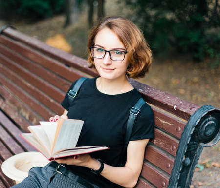 Young attractive woman with short haircut and glasses reads  book while sitting on bench outdoorsの写真素材
