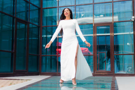Attractive afro woman in a luxurious white dress against a building with blue windowsの写真素材