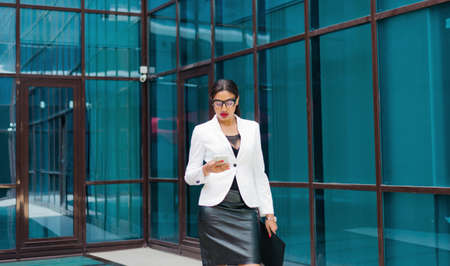 Professional business afro woman in elegant white jacket and skirt with folder with documents in her hands  use smartphone against the background of business center with blue windowsの写真素材