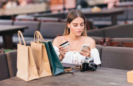 Online shopping. Young woman sits at table with shopping bags and holds in her hands smartphone and bank card in an outdoor cafeの写真素材