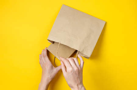 Female hands put a cardboard box in a paper bag on a yellow background. Top view