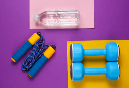 Flat lay style sport concept. Dumbbells, jump rope, bottle of water. Sports equipment on colored...