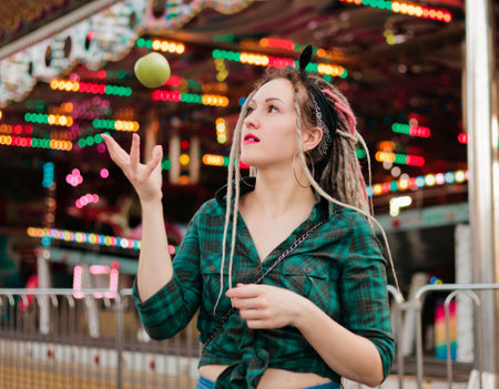 Inordinate young woman with dreadlocks hairstyle and fashionable summer clothes throws apple in amusement park.の写真素材