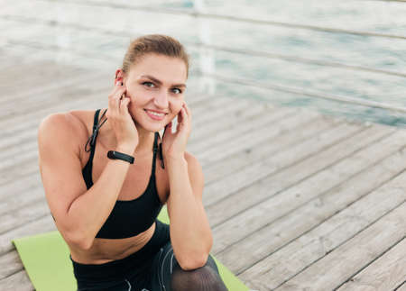 Young fit woman listening to music with earphones while sitting on a mat at wooden terrace on the...