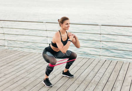 Young sport woman doing squats with rubber band on a beach terrace