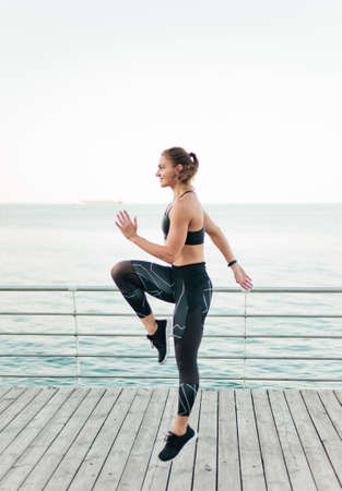 Young fit woman in sportswear doing a high jump on a beach terrace against the sea. Outdoor Sports...