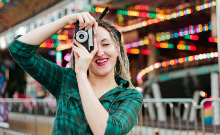 An informal young woman with dreadlocks takes pictures with retro camera in amusement park.