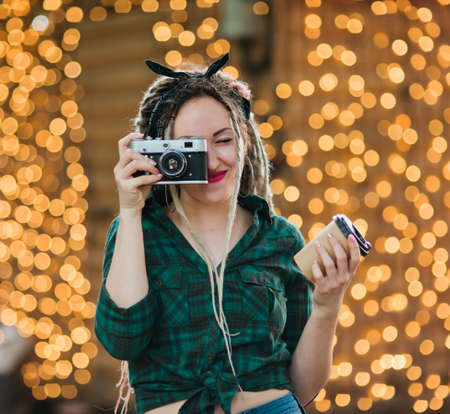 Happy young woman with dreadlocks hairstyle and fashionableand pin-up style clothes takes photo while holding retro camera and coffee cup in his hands against the background of garlandsの写真素材