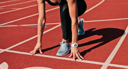 Young runner woman in sportwear getting ready to run sprint at low start on stadium track with red coated at bright sunny day. Crop photoの写真素材