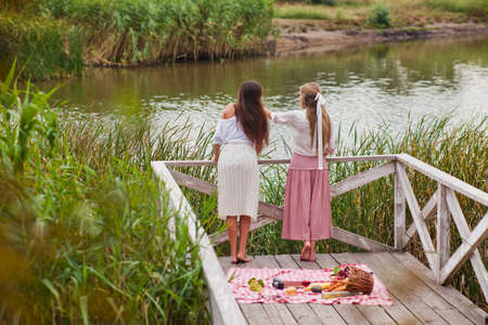 Two cheerful young women have a picnic outdoors on a summer day. Two girlfriends in retro vintage style clothes spend time together on the pond pier.の写真素材
