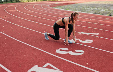Young runner woman in sportwear getting ready to run sprint at low start on stadium track with red...