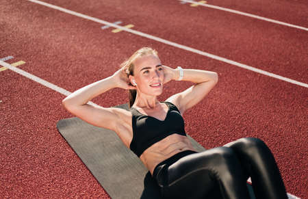 Young fit woman perfoms her body doing twisting exercise for abdominal muscles on mat at stadium track with red coating on bright sunny day. Healthy lifestyleの写真素材