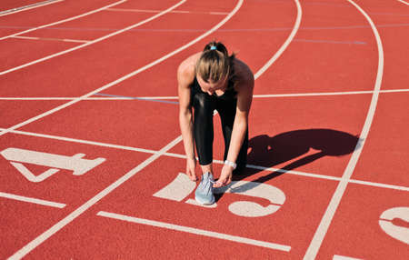 Young sprinter woman in sportswear tying shoelaces before a race on a red-coated stadium track at...