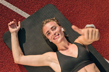 Happy cheerful woman in sportswear lies on  mat at red-coated stadium track and listens to music on...