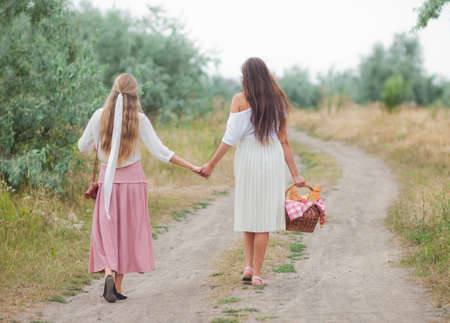 Two cheerful young women in retro style clothes are walking along the landing with a picnic basket and hold hands. Together walk in natureの写真素材
