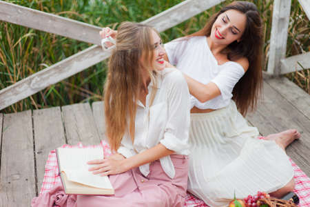 Two cheerful young women have a picnic outdoors on a summer day. Two girlfriends in retro vintage style clothes spend time together on the pond pier.の写真素材