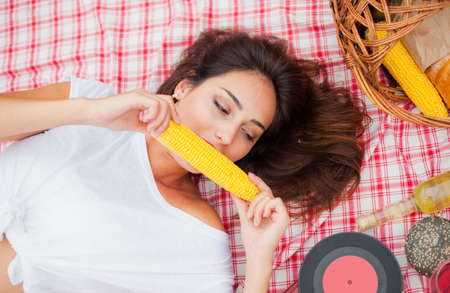 Retro style picnic. Young cheerful blonde woman in vintage clothes eat corn while lies on a picnic...