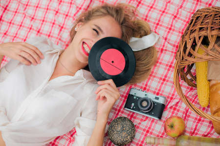 Retro style picnic. Young cheerful blonde woman in vintage clothes holding vinyl record at face while lies on picnic tablecloth outdoors. Top viewの写真素材