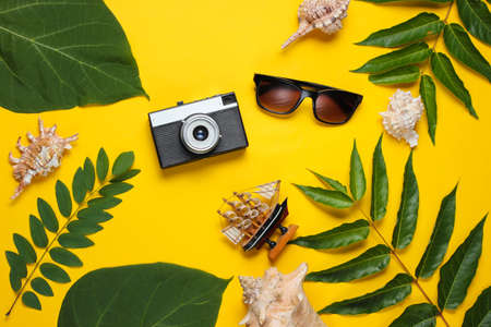 Retro style travel still life. Film camera, sunglasses, seashells, green tropical leaves. Traveler accessories on yellow background. Top viewの写真素材