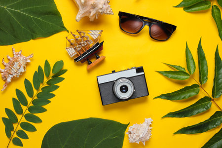 Retro style travel still life. Film camera, sunglasses, seashells, green tropical leaves. Traveler accessories on yellow background. Top viewの写真素材