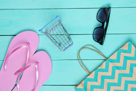 Summertime sale. Shopping concept. Summer beach accessories (flip flops, sunglasses), mini shopping trolley on a blue wooden background. Top view. Flat layの写真素材