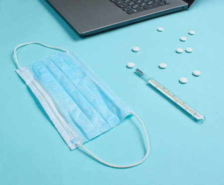 Medical background. Workspace of a modern doctor. Laptop, pills, gauze mask, thermometer on a blue background.の写真素材