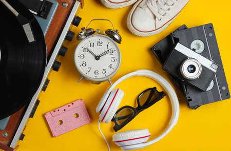 Flat lay retro 80s pop culture objects. Vinyl player, headphones, audio cassette, video tapes, alarm clock, film camera, sneakers on yellow background. Top viewの写真素材