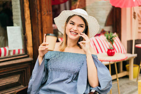 Young fashionable woman in a hat and dress sits at outdoor cafe and drinks coffee while talking on phoneの写真素材