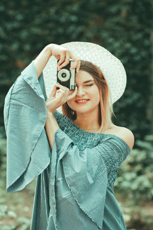 Vintage portrait. Young spectacular woman in dress and hat takes pictures with retro camera on blurred outdoor backgroundの写真素材