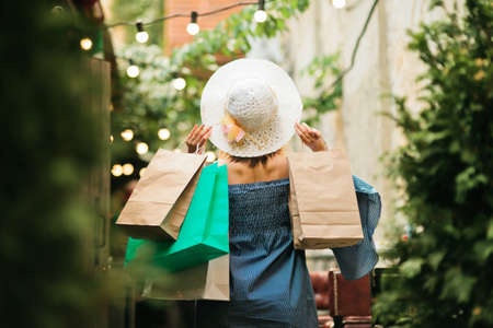 Happy shopping concept. Young attractive woman in a hat and dress holds shopping bags in her hands while walking along the street. Back viewの写真素材